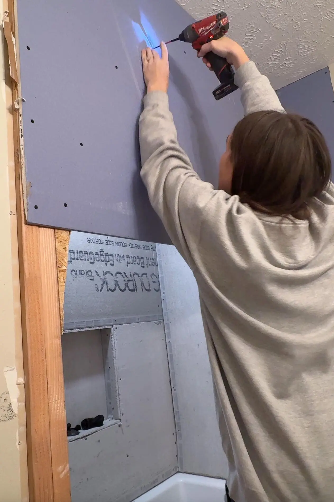 A woman using a red power drill to secure a large purple drywall sheet onto a bathroom wall. She stands on the edge of the bathtub, ensuring the drywall is firmly attached to the wooden frame.